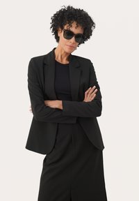 Blazer nero su un top nero, abbinato a una gonna nera. Capelli ricci e occhiali da sole scuri, braccia incrociate, sfondo neutro.