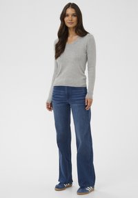 Femme aux longs cheveux noirs portant un pull V-neck gris clair, un jean bleu droit et des baskets bleues, se tenant devant un fond uni.