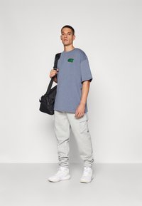 Grå cargobukser og en blå oversized t-shirt med et grønt logo. Personen bærer en sort taske og har hvide træningssko på.