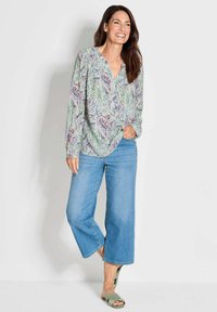Leichte Bluse mit langen Ärmeln und mehrfarbigem Blumenmuster, kombiniert mit weit geschnittenen Jeans und grünen Sandalen mit offenen Zehen.