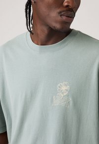 Πράσινο βαμβακερό μπλουζάκι με μικρό, κεντητό λουλουδένιο σχέδιο σε λευκό στο στήθος. Κοντά μανίκια και στρογγυλή λαιμόκοψη.