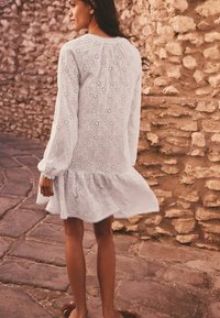 Robe blanche en broderie anglaise avec des manches longues, un ourlet en forme de scallop, des motifs floraux et une coupe ample, portée avec des chaussures marron à enfiler.