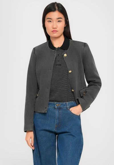 Vrouw in een donkergrijze jasje met knopen over een zwart-wit gestreept shirt en een blauwe jeans, staande met één hand in de zak tegen een witte achtergrond.