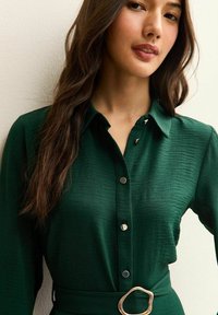 Zielona koszula z guzikami, wykonana z teksturowanej tkaniny, z kołnierzykiem i ozdobnym złotym zapięciem na pasku w talii. Srebrne guziki zdobią przód.
