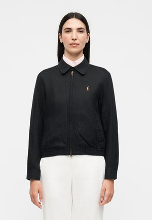 Vrouw met stijl donker haar, gekleed in een zwarte jas met rits over een lichtroze overhemd met kraag en een witte broek, staand tegen een effen achtergrond.