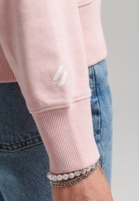Sudadera rosa claro con puños acanalados, que presenta un logo blanco en la manga. Combinada con jeans de mezclilla azules y pulseras en capas.