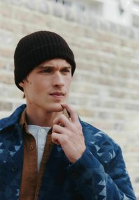 Modrý vzorovaný blejzer s trojuholníkovými vzormi, hnedá košeľa a biela štruktúrovaná henley. Nosič má čiernu rebrovanú čiapku a dotýka sa brady.