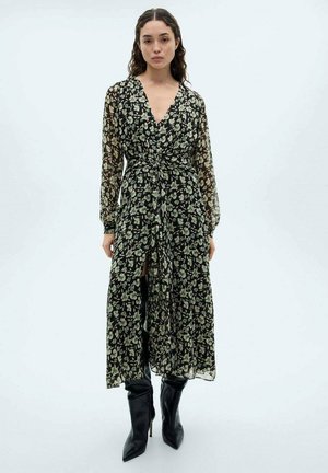 Robe florale noire avec de longues manches transparentes, un décolleté en V et une taille cintrée. Présente des motifs floraux verts et crème ainsi qu'une fente.
