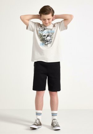 Dreng iført en t-shirt med grafisk kranium, sorte shorts, stribede sokker og grå sneakers, stående med hænderne bag hovedet foran en hvid baggrund.