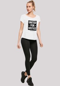 Balta trumpa rankovėmis marškiniai su tekstu "UNITED FOR JUSTICE" juoda spalva, derinami su juodomis legginso kelnėmis ir juodais sportiniais batais.