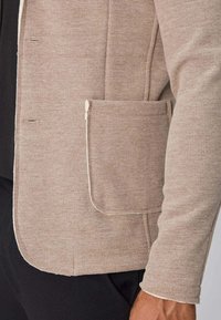 Manche de veste beige côtelée et poche à rabat sur un pantalon noir, montrant des détails de texture et de couture.