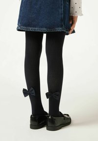 Le calze a coste blu notte presentano due fiocchi abbinati alle caviglie, abbinate a scarpe in pelle verniciata nera e una sovrapposizione di gonna di jeans.