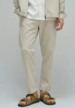 Hombre con pantalones beige, camisa blanca, chaqueta ligera y sandalias marrones con hebillas, de pie frente a un fondo liso.