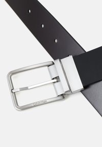 Cintura in pelle nera con fibbia in metallo argentato; caratteristiche includono una texture liscia e diversi fori per la regolazione. "Calvin Klein" inciso sulla fibbia.