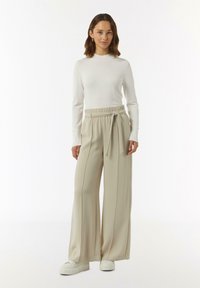 Beige wijde broek met een strik taille, gemaakt van een gladde stof. Gecoordineerd met een aansluitend wit shirt met lange mouwen en witte platform sneakers.