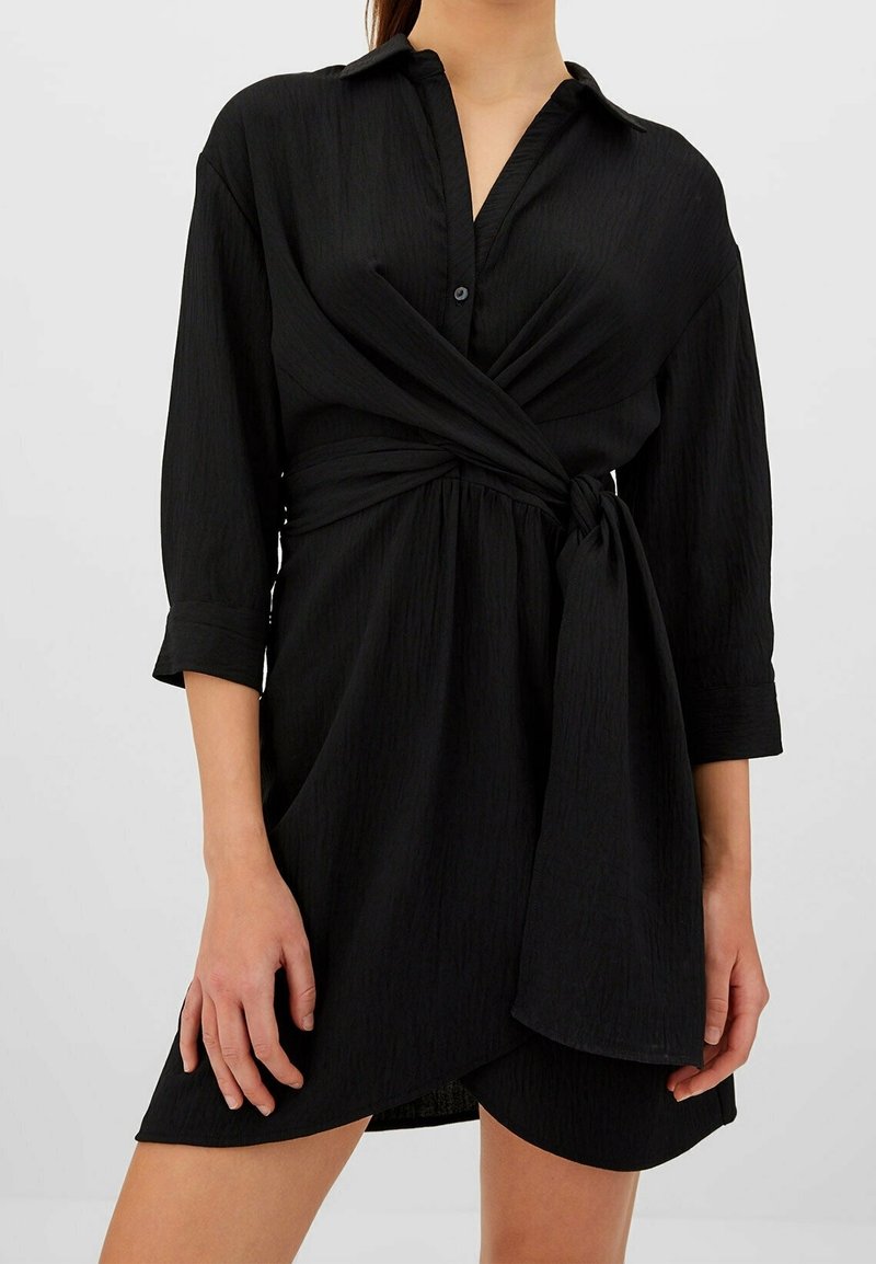 Femme portant une robe portefeuille noire à manches longues, col, attache latérale et tissu texturé, debout devant un fond clair et uni.