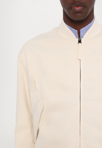 Veste bomber crème avec une texture lisse, fermeture éclair sur le devant, accessoires noirs et une poche latérale. Design avec col orné d'un tissu à carreaux bleu.