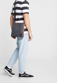 Šedá crossbody taška z látky s oranžovými akcenty, čtvercový tvar, nastavitelný popruh, viditelná nášivka s logem, nošená s světle modrými džíny a černými teniskami.