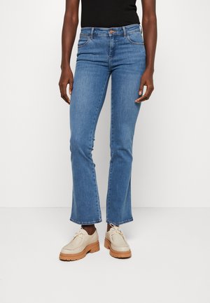 Personne portant un jean bleu taille normale à jambes droites et des chaussures beiges à lacets avec semelles épaisses, debout sur un fond blanc uni.