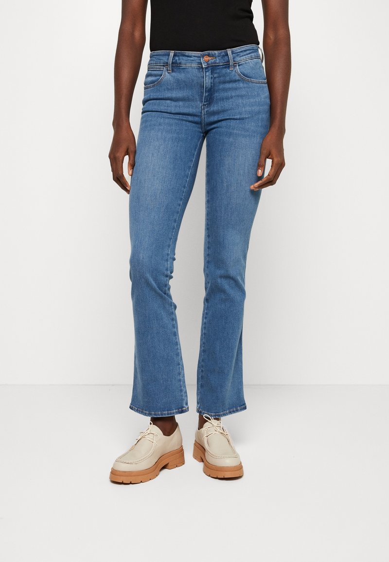 Personne portant un jean bleu taille normale à jambes droites et des chaussures beiges à lacets avec semelles épaisses, debout sur un fond blanc uni.