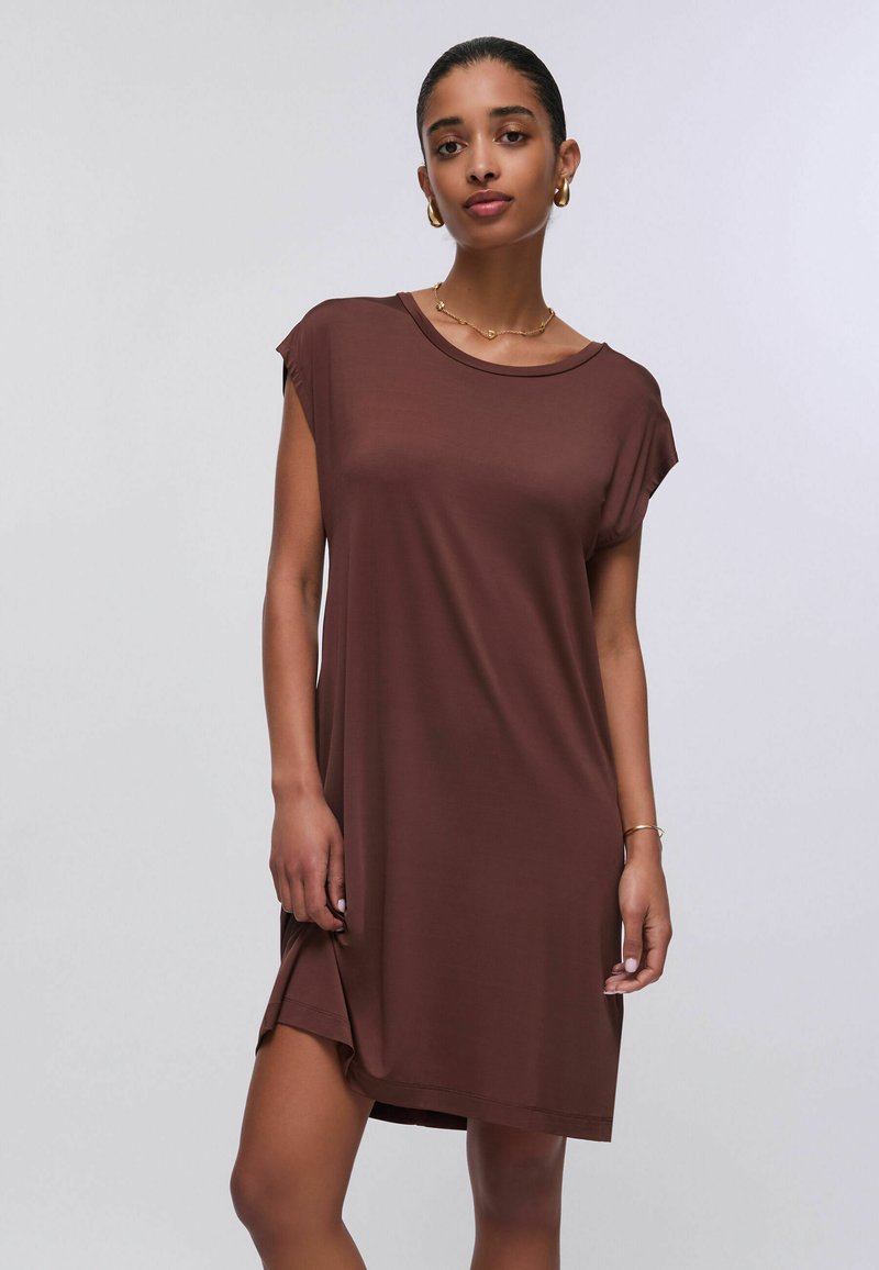Femme portant une robe marron à manches courtes avec un col rond, des boucles d'oreilles créoles dorées, un collier et un bracelet sur un fond clair uni.