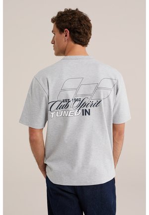 Jeune homme de dos portant un t-shirt gris clair avec un texte en noir et blanc « Club Spirit Tuned In EST. 1962 » et un motif géométrique dans le dos.