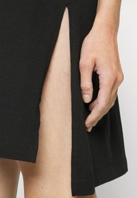 Robe noire avec une fente sur le côté, montrée avec une main posée sur le tissu. Texture douce et design simple avec des coutures détaillées.