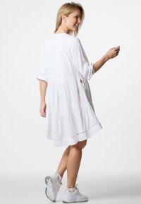 Weißes, knielanges Kleid mit langen Ärmeln, das einen gestuften Rock und eine dezente Textur aufweist. Kombiniert mit weißen Sportschuhen.