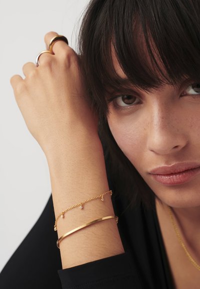 Zlaté šperky na zápästí obsahujú jemný retiazkový náramok s malými príveskami a širší plochý náramok. Pokožka má hladkú, svetlú textúru.