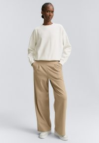 Ženska stoji z rokami v žepih, oblečena v belo dolge rokave, bež široke hlače in bele tene na svetlo sivi ozadju.