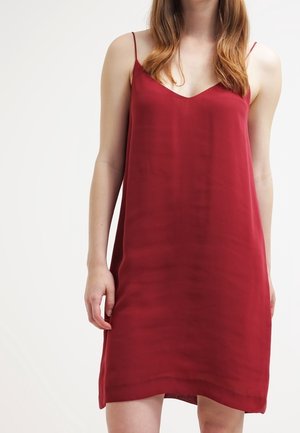 Femme portant une robe nuisette rouge foncé sans manches avec de fines bretelles, debout devant un fond blanc uni.
