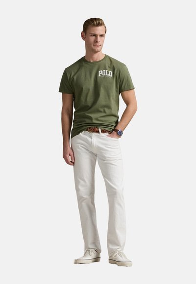 Camiseta de algodón verde oliva con el logo de 'Polo Ralph Lauren', combinada con pantalones de algodón blancos y zapatos de color claro, en posición de pie.