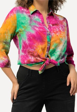 Multifarvet tie-dye skjorte med knapper, bundet ved taljen, båret med sorte højttaljede bukser, 3/4-ærmer og krøllet hår synligt.