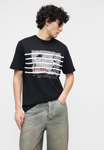 Jeune homme aux cheveux bouclés portant un t-shirt noir avec un motif graphique et un jean clair, posant avec une main dans la poche et regardant sur le côté.