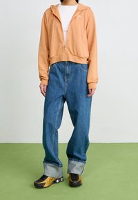 Orange zip-up hoodie med en framficka, tillsammans med lösa blå denimbyxor med uppvikta nederkanter. Svart och gula skor fulländar looken.