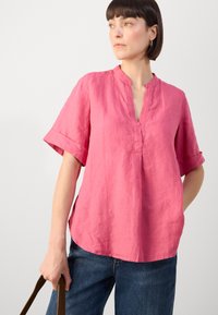 Blusa corta a maniche corte in lino rosa con scollatura a V e risvolti, caratterizzata da una vestibilità rilassata e fessure laterali. Indossata con jeans blu scuro.