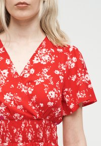 Γυναίκα με ανοιχτόξανθα μαλλιά που φοράει κόκκινο φόρεμα με λουλούδια, V λαιμόκοψη, κοντά μανίκια που πέφτουν απαλά και μέση με σούρες.