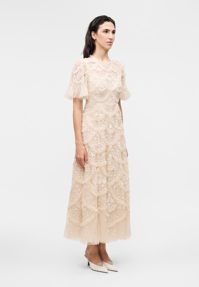 Γυναίκα στέκεται φορώντας μια μπεζ, κεντημένη, γόνατο-μακρύ φόρεμα με φουσκωτά διάφανα μανίκια και μυτερά ψηλοτάκουνα παπούτσια.