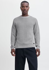 Pull en tricot côtelé gris avec des manches longues, un col rond et un ourlet ajusté. Présente des détails subtils et une étiquette en bas à gauche.