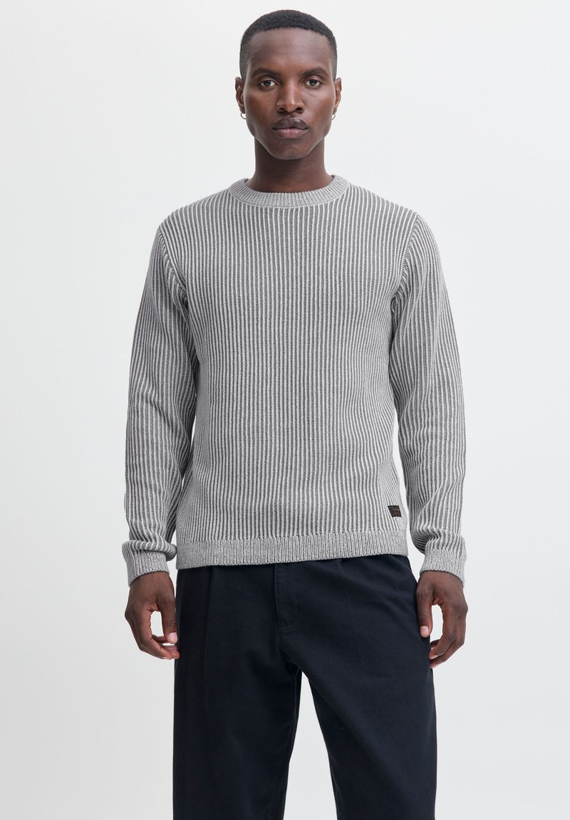 Pull en tricot côtelé gris avec des manches longues, un col rond et un ourlet ajusté. Présente des détails subtils et une étiquette en bas à gauche.