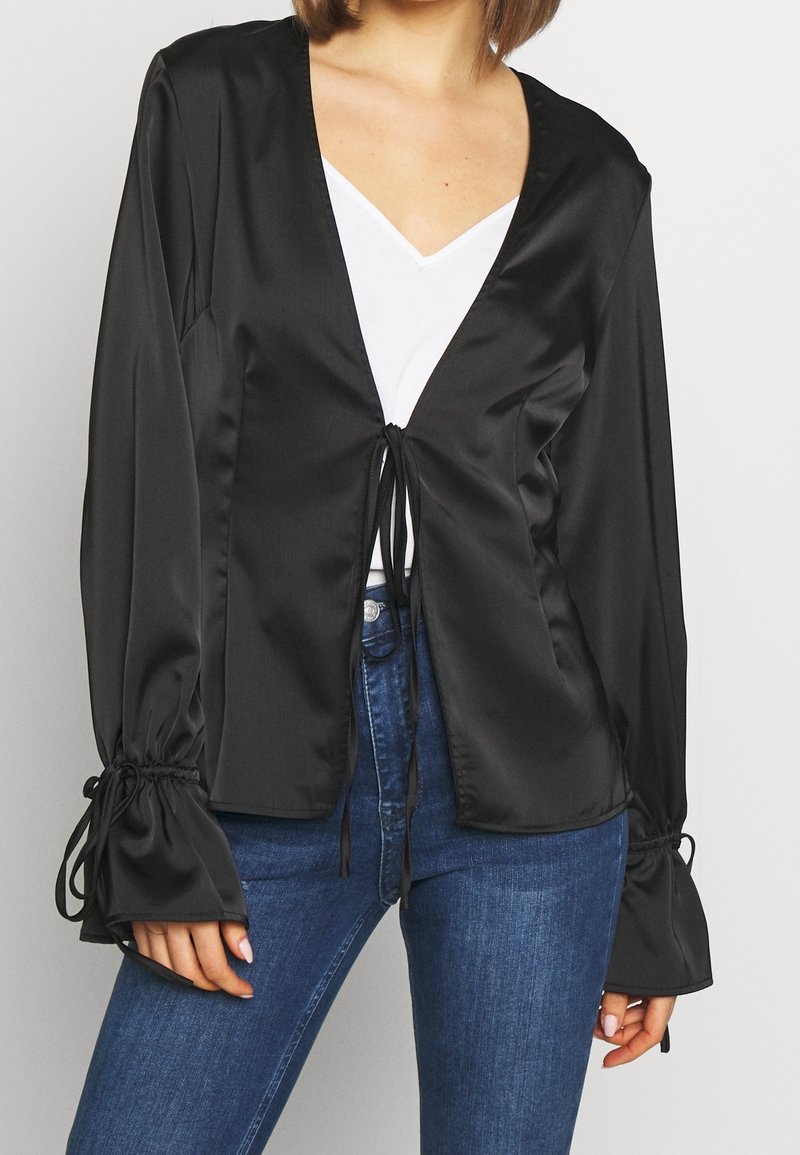 Blouse en satin noir avec un décolleté en V, fermeture à nouer à la taille et manches évasées avec des détails froncés. Portée sur un top blanc.