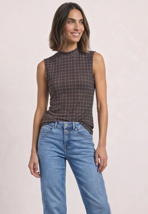 Femme aux cheveux bruns mi-longs portant un top marron à carreaux sans manches et un jean bleu clair taille haute, souriante et regardant vers la droite.