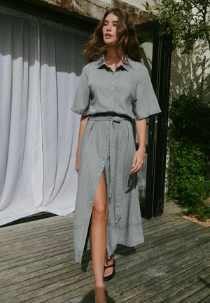 Nő szürke, gombos ingruhában, rövid ujjakkal és övvel, fa teraszon áll, egy ajtó mellett, közelben növényekkel.