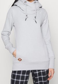 Ljusgrå huvtröja med hög krage, justerbara snören och en frontficka. Har en brun logotypapplikation på fållen. Mjuk tygkvalitet.