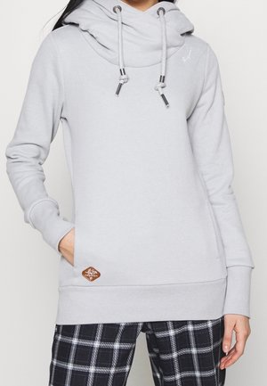 Sudadera gris claro con cuello de embudo, cordones ajustables y un bolsillo frontal. Presenta un parche de logo marrón en el dobladillo. Textura de tela suave.