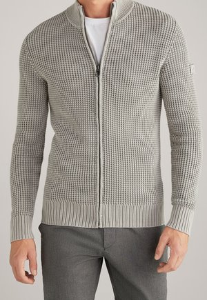 Homme portant un pull zippé gris clair texturé sur une chemise blanche et un pantalon gris, debout avec les bras détendus.
