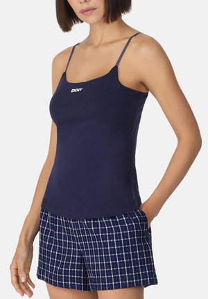Donna che indossa un top a spalline sottili DKNY blu navy e pantaloncini a quadri blu, in piedi con una mano in tasca su sfondo bianco.