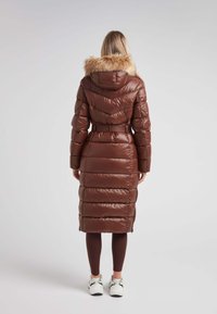 Brun vadderad jacka med en foderhuva av päls, midja med bälte, lång längd och quiltat mönster. Bärs över bruna leggings och vita sneakers.