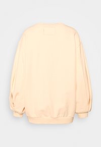 Sweat-shirt pêche clair à coupe décontractée, manches longues bouffantes, encolure ronde, et une petite étiquette en tissu sur le haut du dos. Texture lisse.