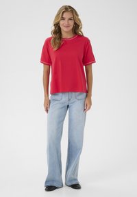 T-shirt rouge à manches courtes avec un col rond et des bordures blanches, associé à un jean évasé bleu clair et des chaussures noires.